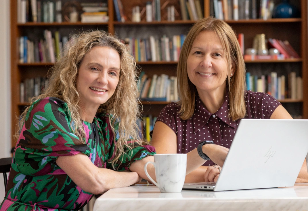 Kristy McGrath and Diane Konstantinou sitting at a desk smiling for photo