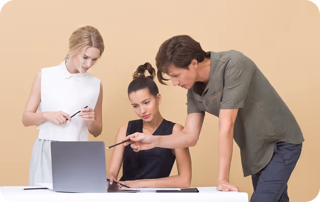 Three people collaborating over a laptop at a white desk with a beige background.