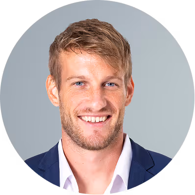 Smiling man with light brown hair and blue eyes wearing a white shirt and navy blazer against a gray background.