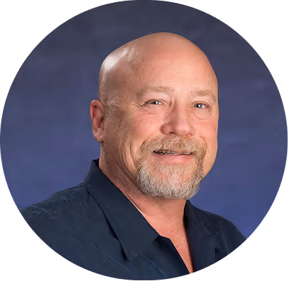 Smiling bald man with a goatee wearing a dark blue shirt against a blue gradient background.
