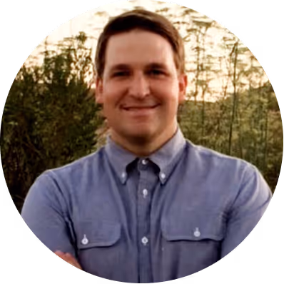 Smiling man with short brown hair wearing a blue button-up shirt, standing outdoors with greenery in the background.