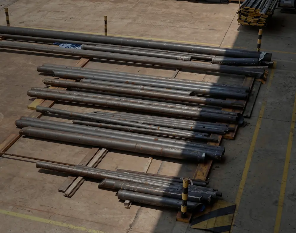 Stacks of long metal pipes arranged on wooden pallets in an outdoor industrial area with concrete flooring.