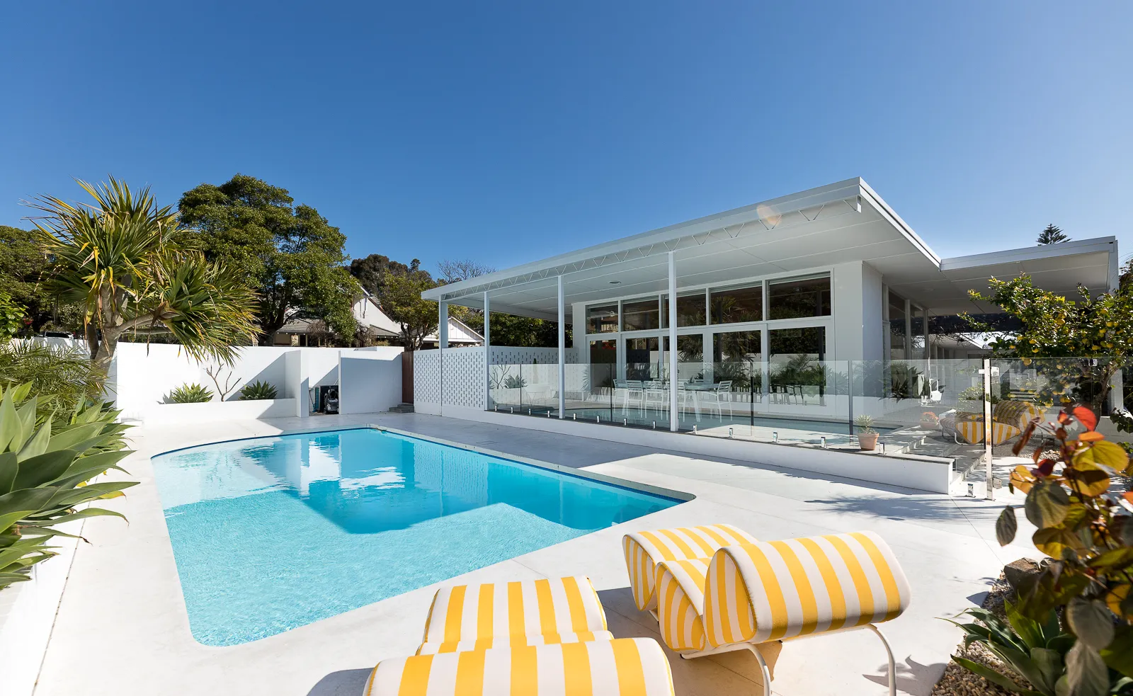 Modern white house with glass-paneled patio and blue swimming pool under clear blue sky, featuring yellow and white striped loungers.