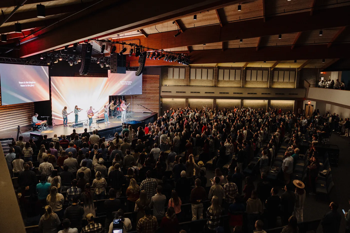 Indoor service with a large congregation standing and a band leading worship on stage with two large screens displaying song lyrics.