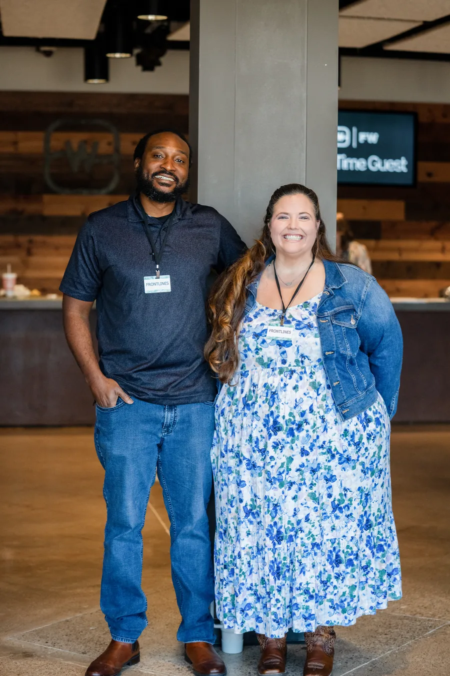 Smiling man wearing a black polo shirt and blue jeans stands next to a smiling woman in a floral dress with a denim jacket; both wear 'FRONTLINES' badges.