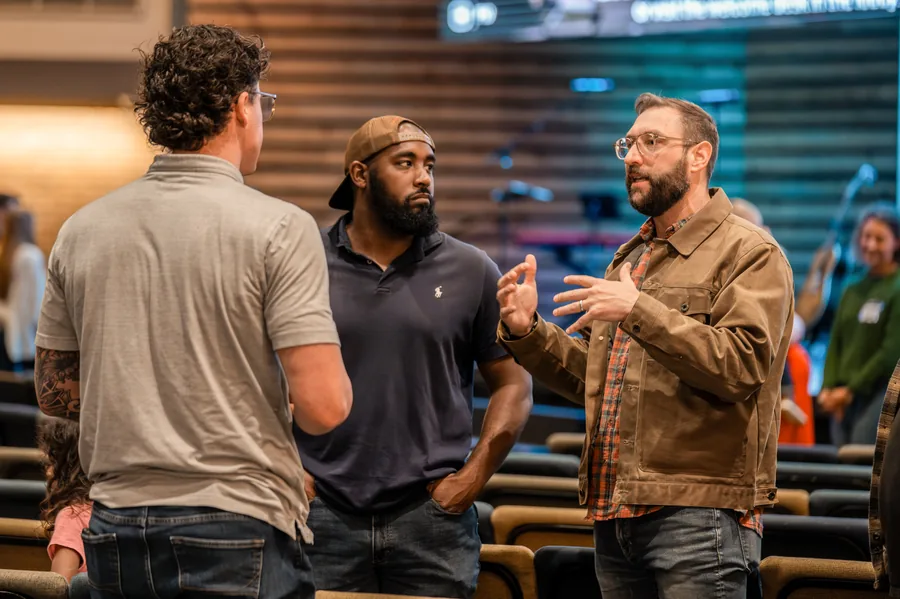 Three men standing and engaged in conversation in an indoor setting with rows of seats in the background.