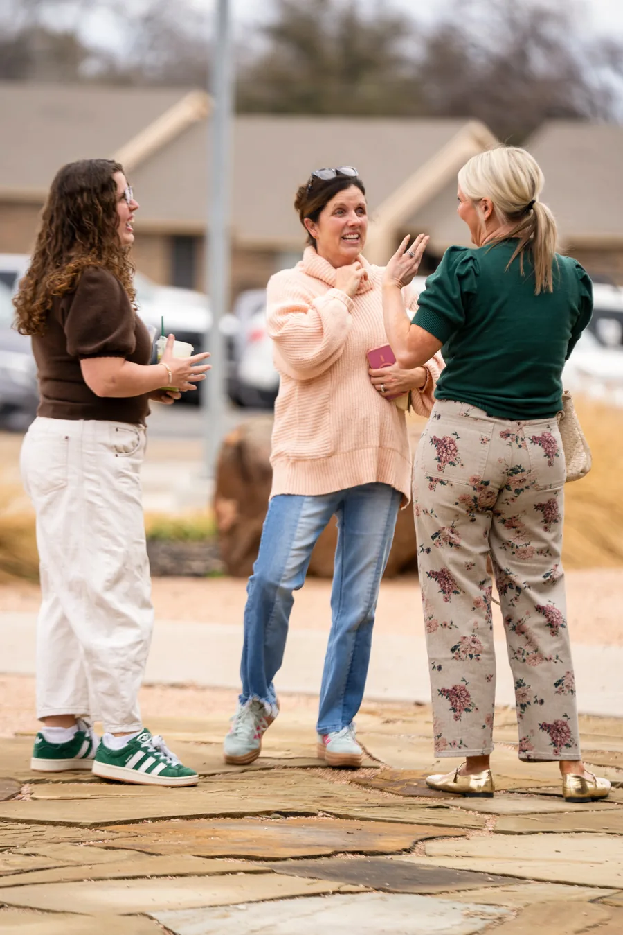 Three women standing outdoors engaged in lively conversation, one holding a drink cup.