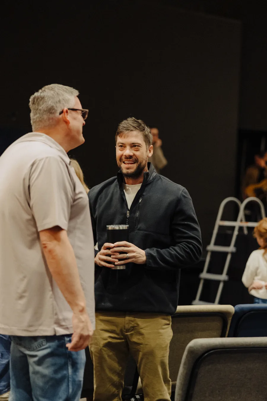 Two men engaged in conversation indoors, one holding a tumbler, with a ladder and chairs in the background.