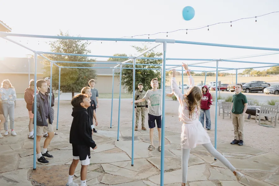 Group of children playing with a blue balloon outdoors under a metal frame structure on a stone patio.