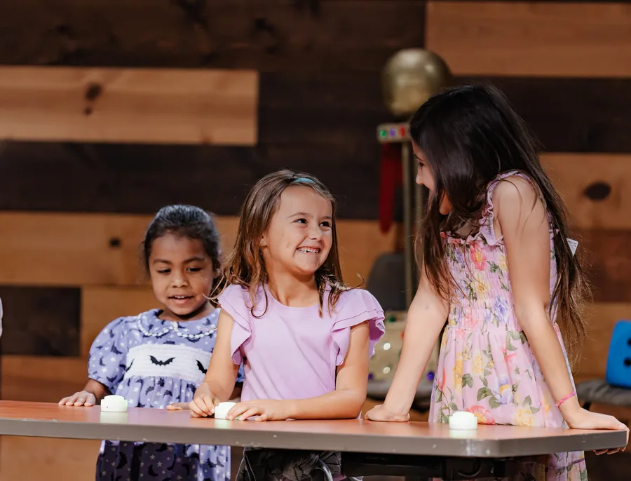Three young girls standing and sitting at a table, smiling and interacting in a warmly lit room with wooden walls.