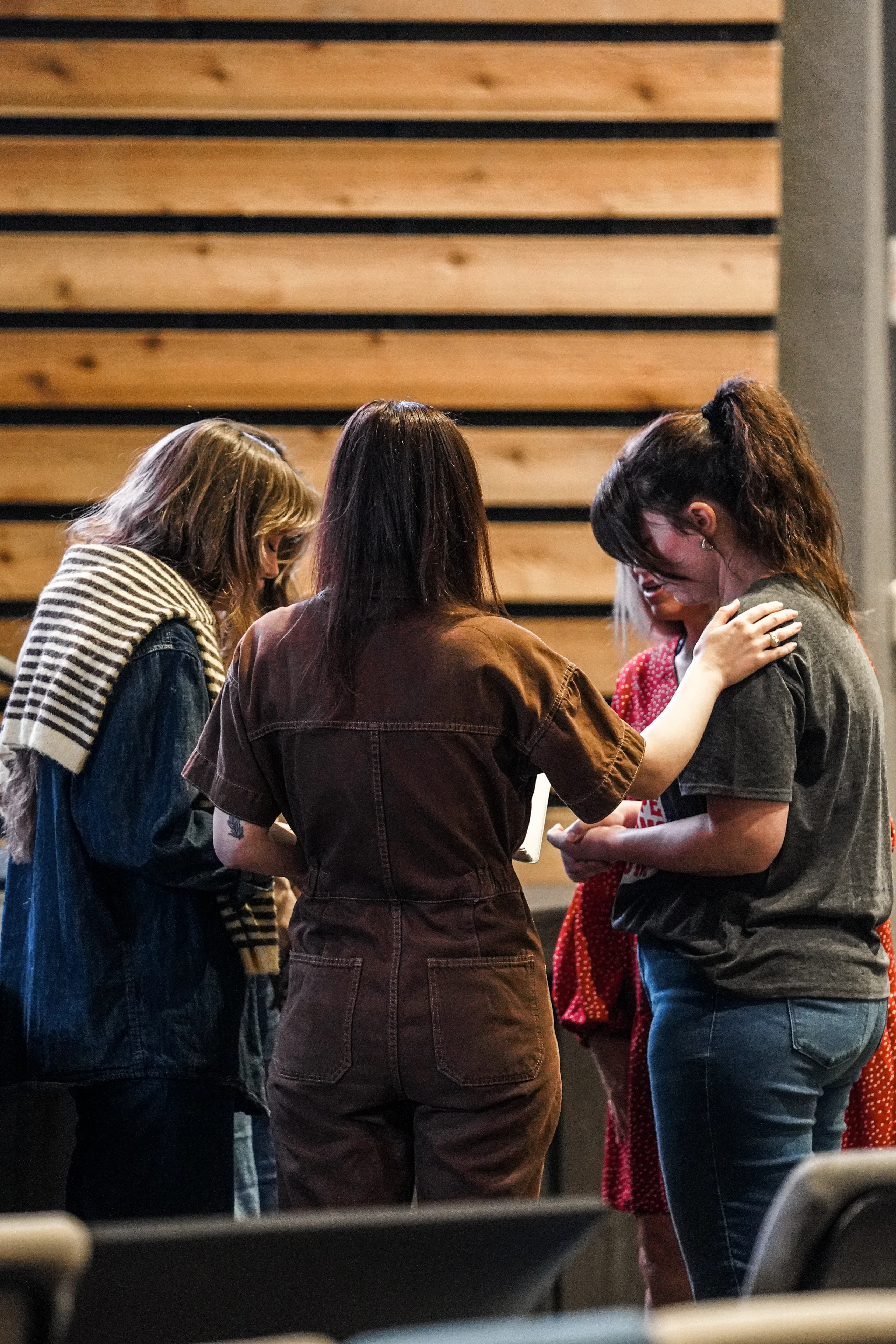 Four women standing close together indoors in a supportive circle, one woman gently touching another's shoulder.