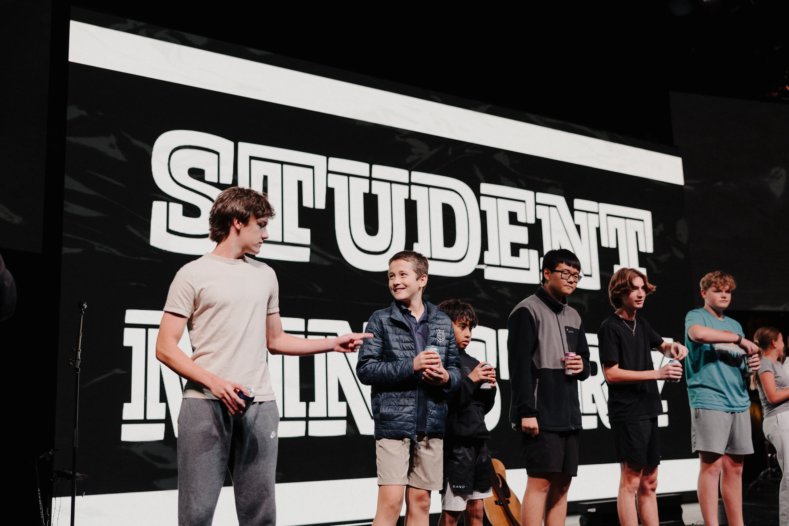 Six young students standing on stage holding cans, with a large screen behind them displaying the word STUDENT.