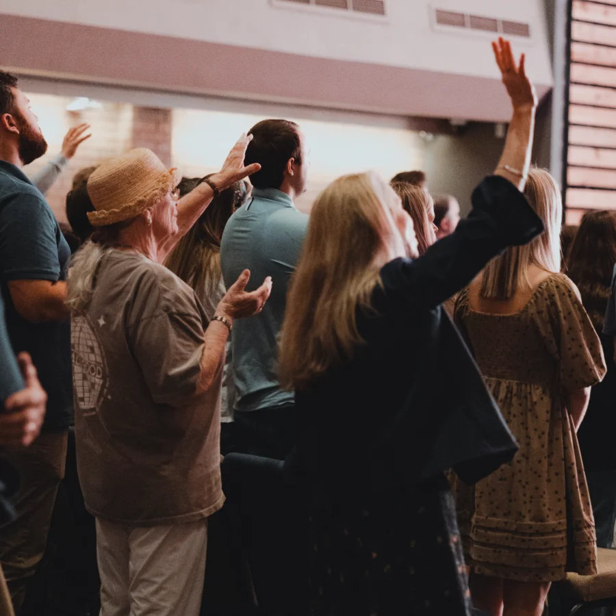 Group of people standing indoors with hands raised, appearing to participate in a worship gathering.