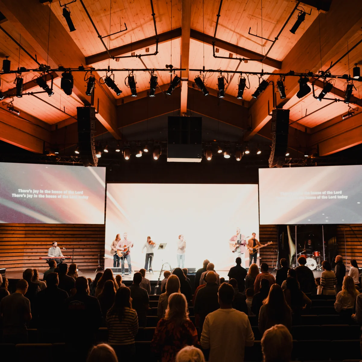 Congregation stands in a modern church with a worship band on stage under wooden ceiling and two projection screens.