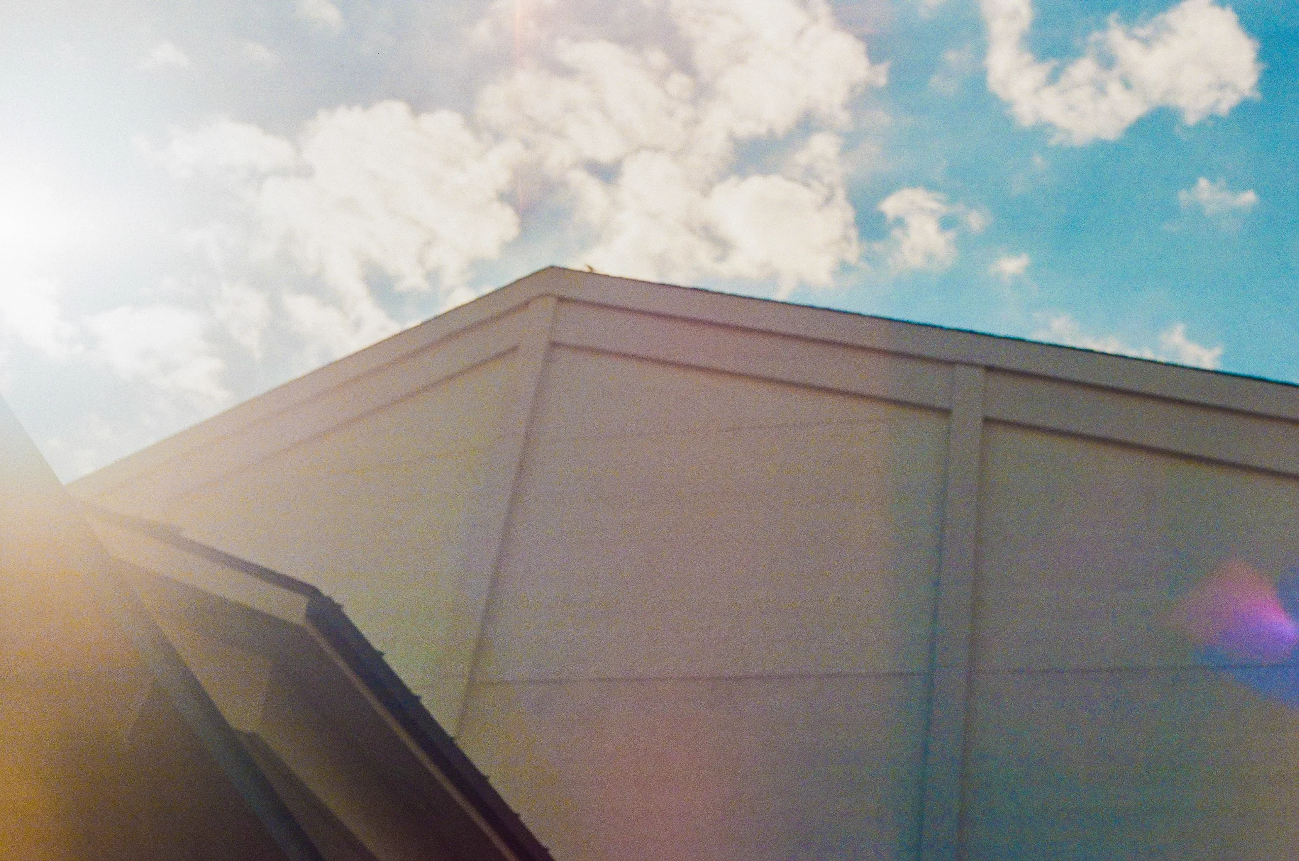 Corner of a beige building against a blue sky with white clouds and sunlight flare.