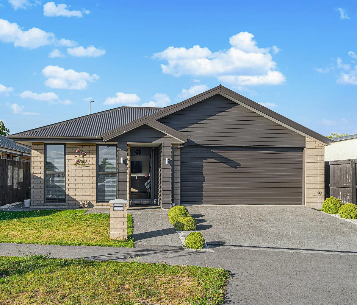 Modern single-story house with a gray roof, dark garage door, brick walls, and a small lawn with bushes in front.