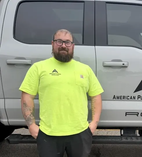 Bearded man with tattoos wearing glasses and a yellow American Plumber t-shirt standing in front of a white truck.