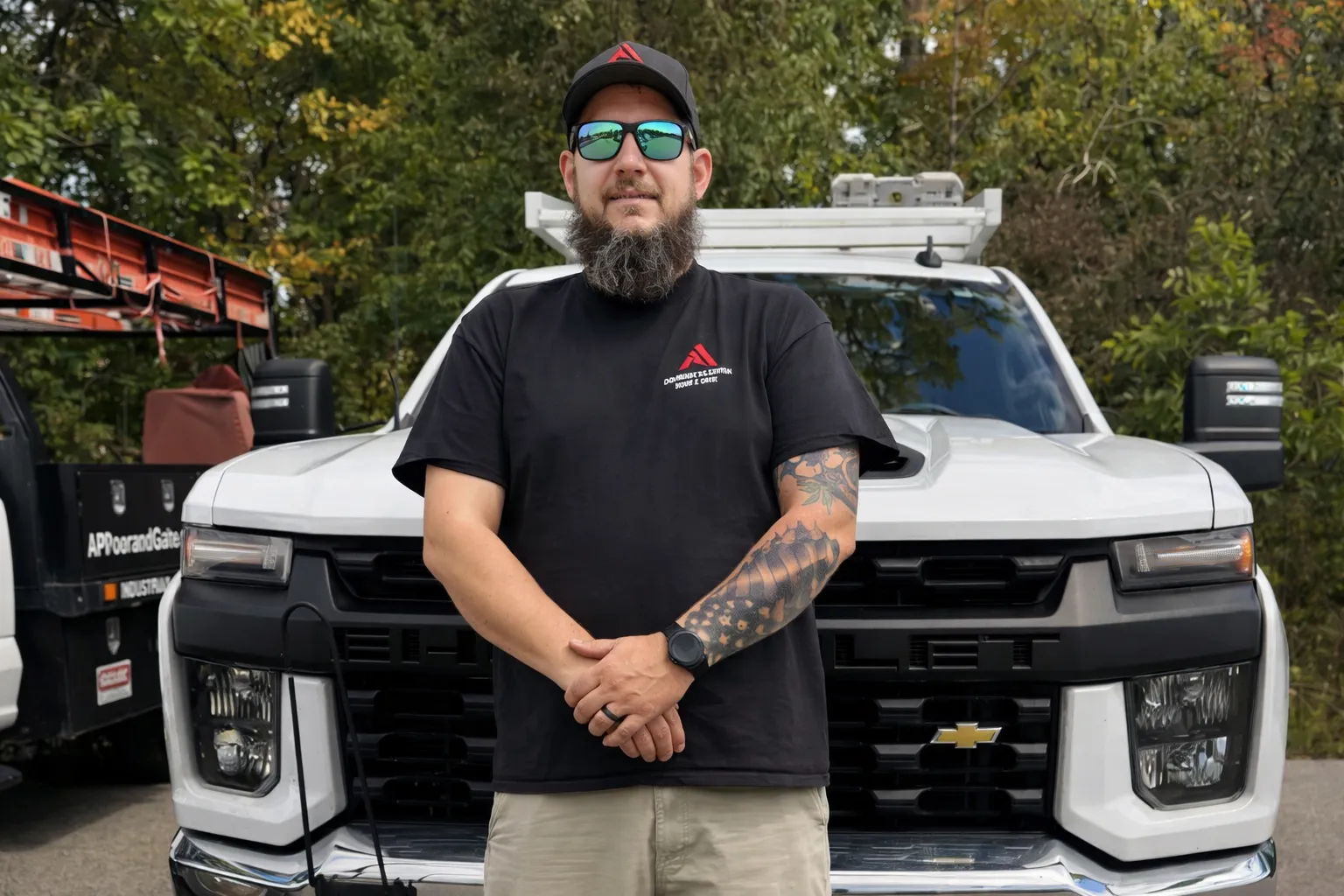 Man with sunglasses, beard, and tattoos standing in front of a white Chevrolet truck.