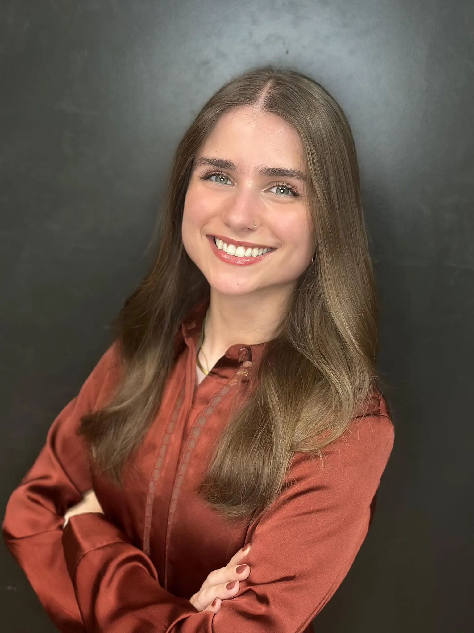 Smiling young woman with long light brown hair wearing a rust-colored blouse against a dark background.