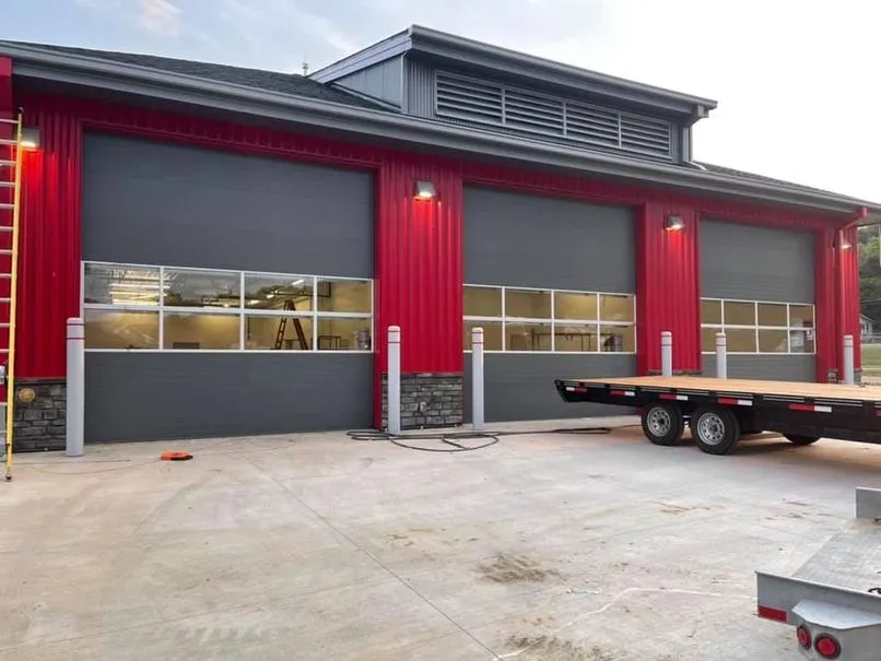 Building with red siding and three large closed gray garage doors with windows, a trailer parked on the right, and concrete pavement in front.