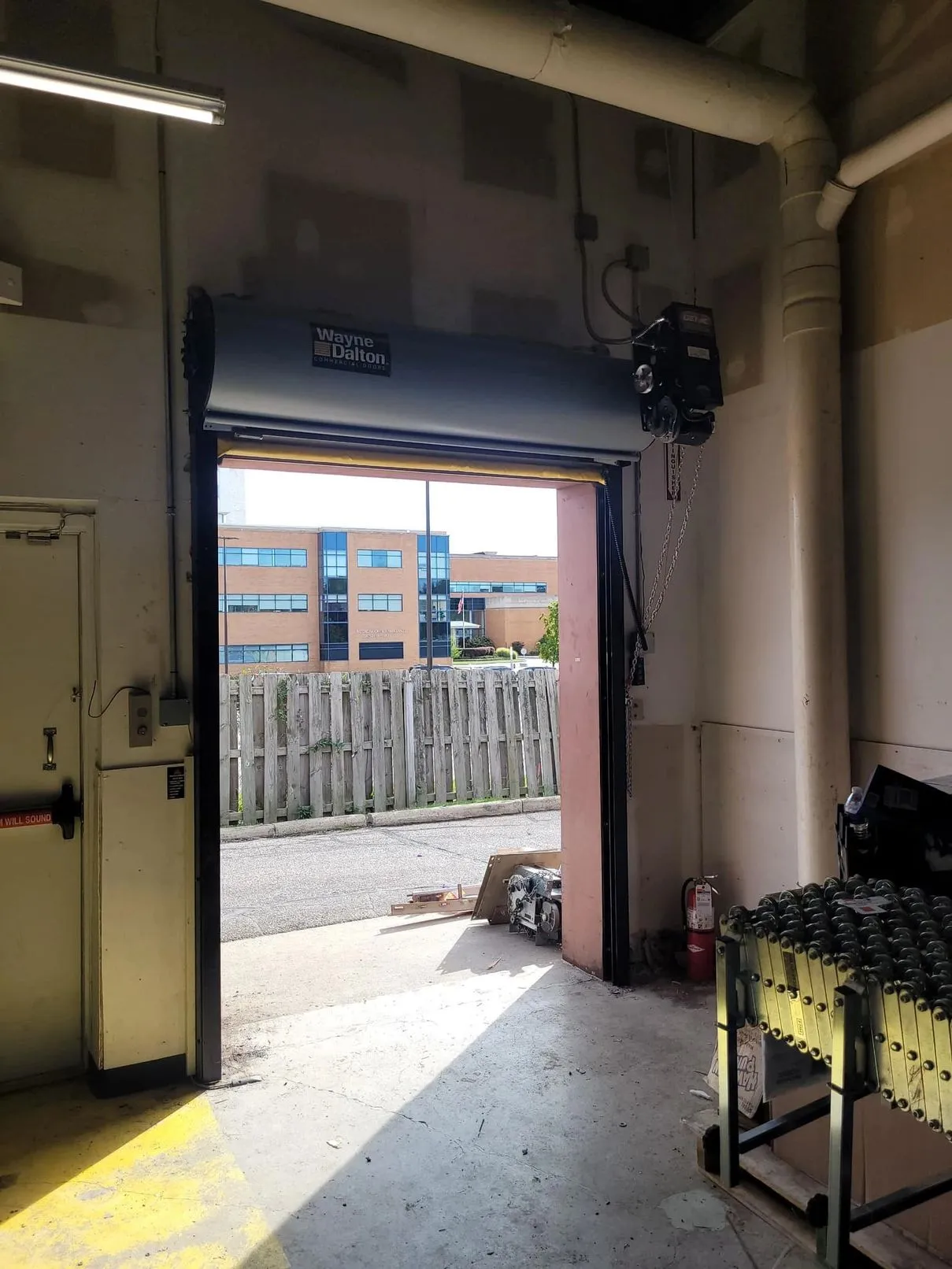 Industrial warehouse interior showing a black pedestrian door within a larger gray commercial rolling door, with lockers and storage along the left wall and an EXIT sign above.