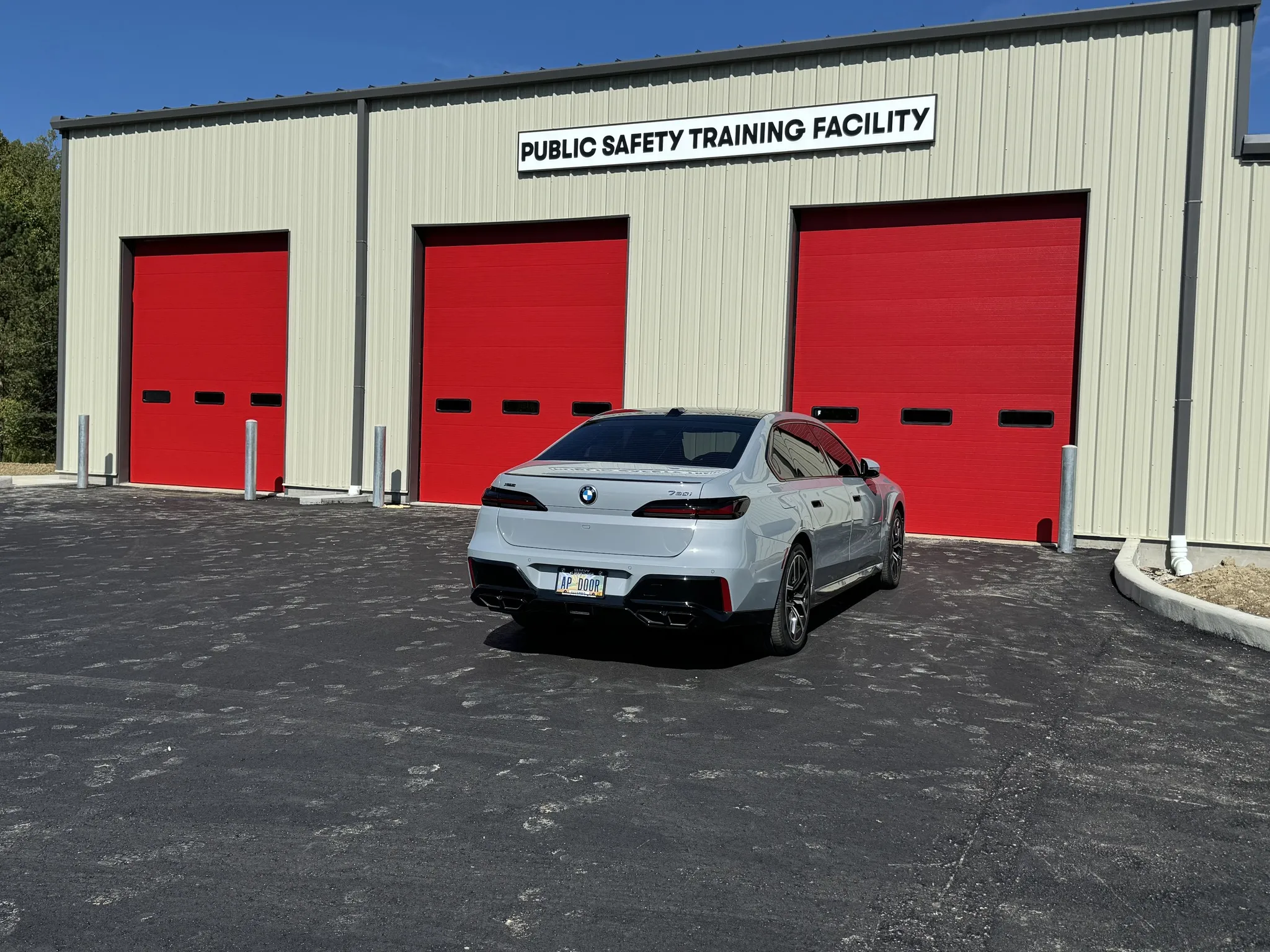 Silver BMW 7 Series sedan parked in front of a beige building with three large red garage doors and a sign reading Public Safety Training Facility.