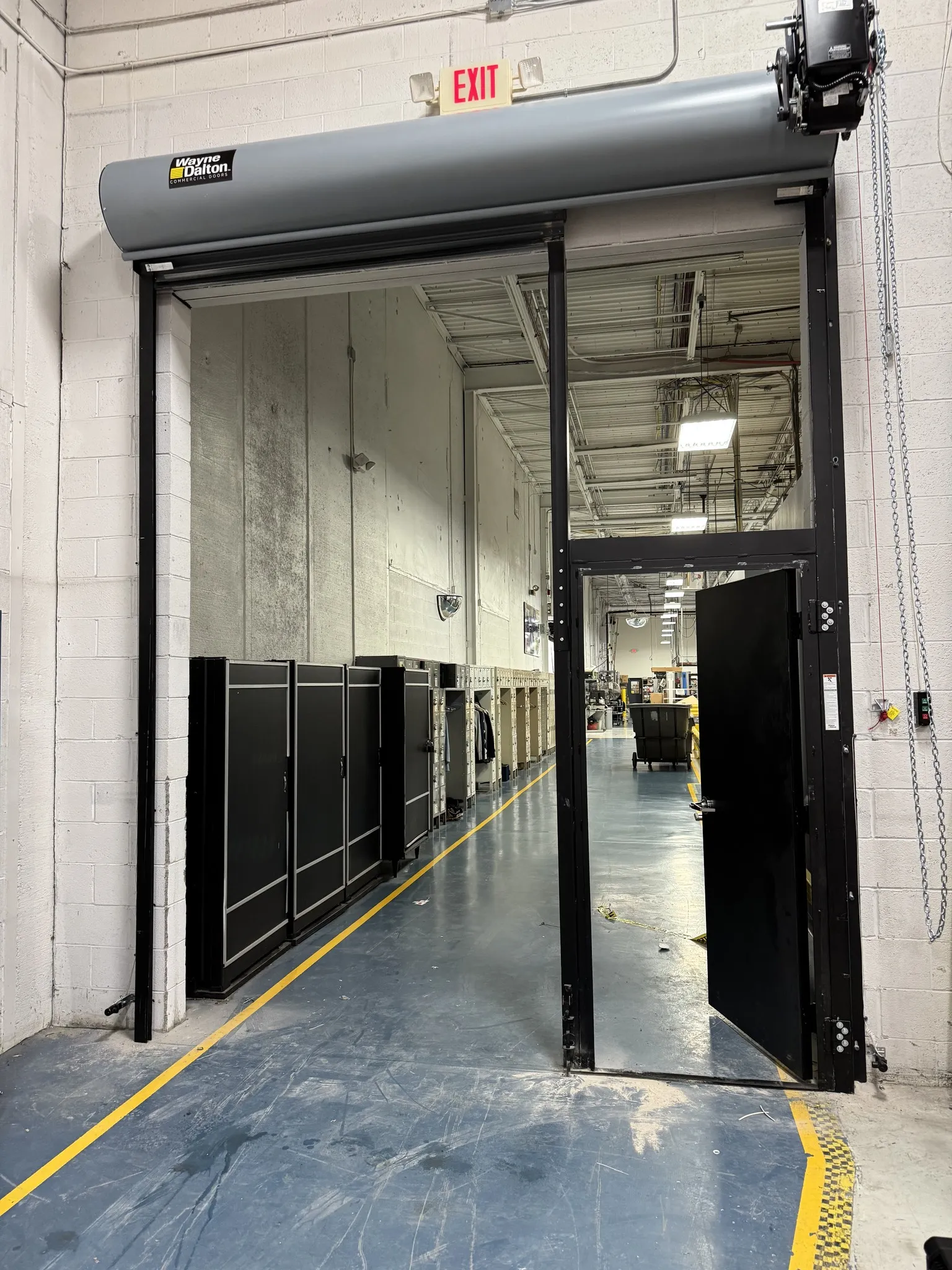 Interior view of an industrial garage with a partially open Wayne Dalton roll-up door, showing a wooden fence and a brick building outside.