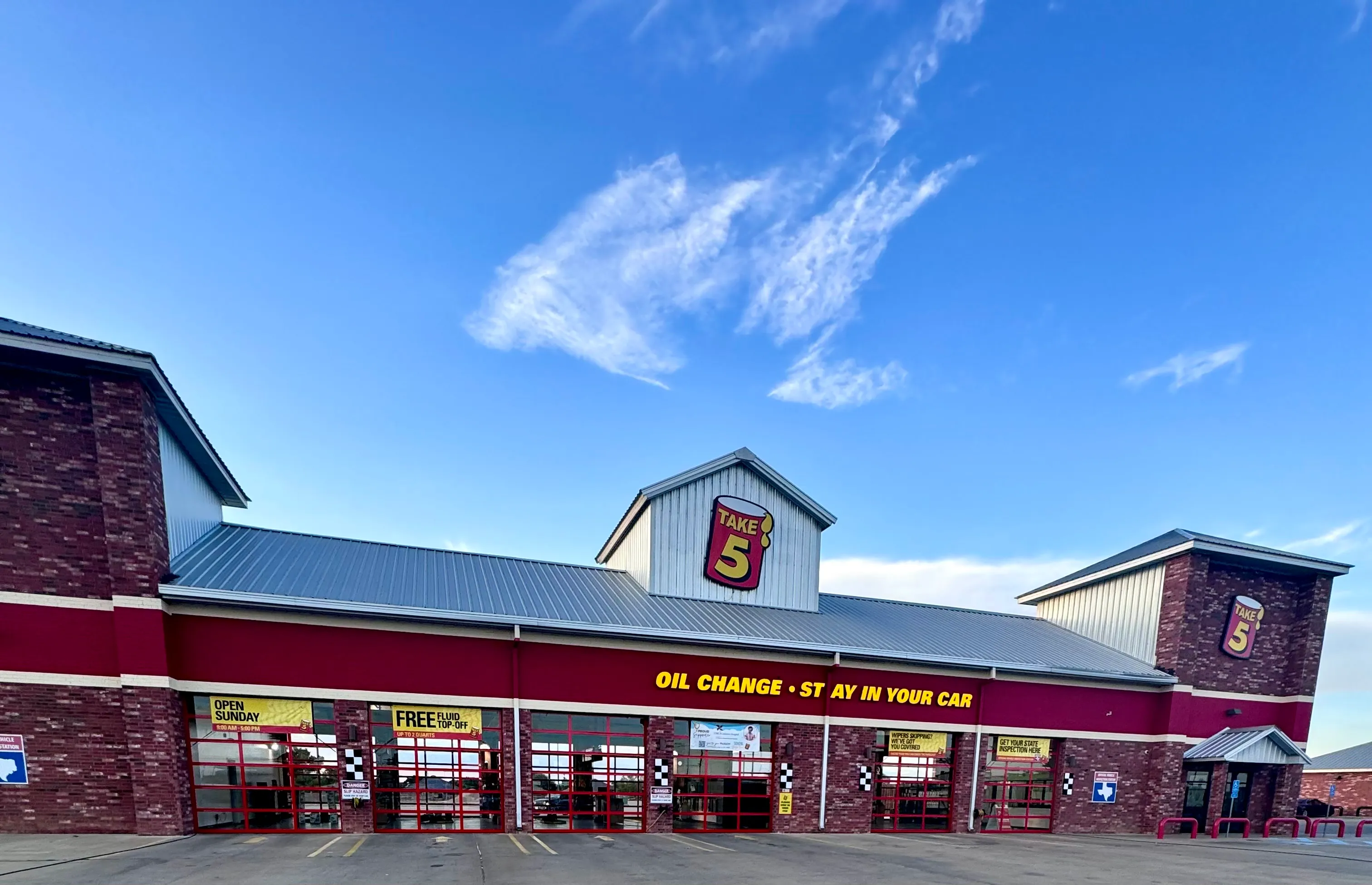 Building with red siding and three large closed gray garage doors with windows, a trailer parked on the right, and concrete pavement in front.