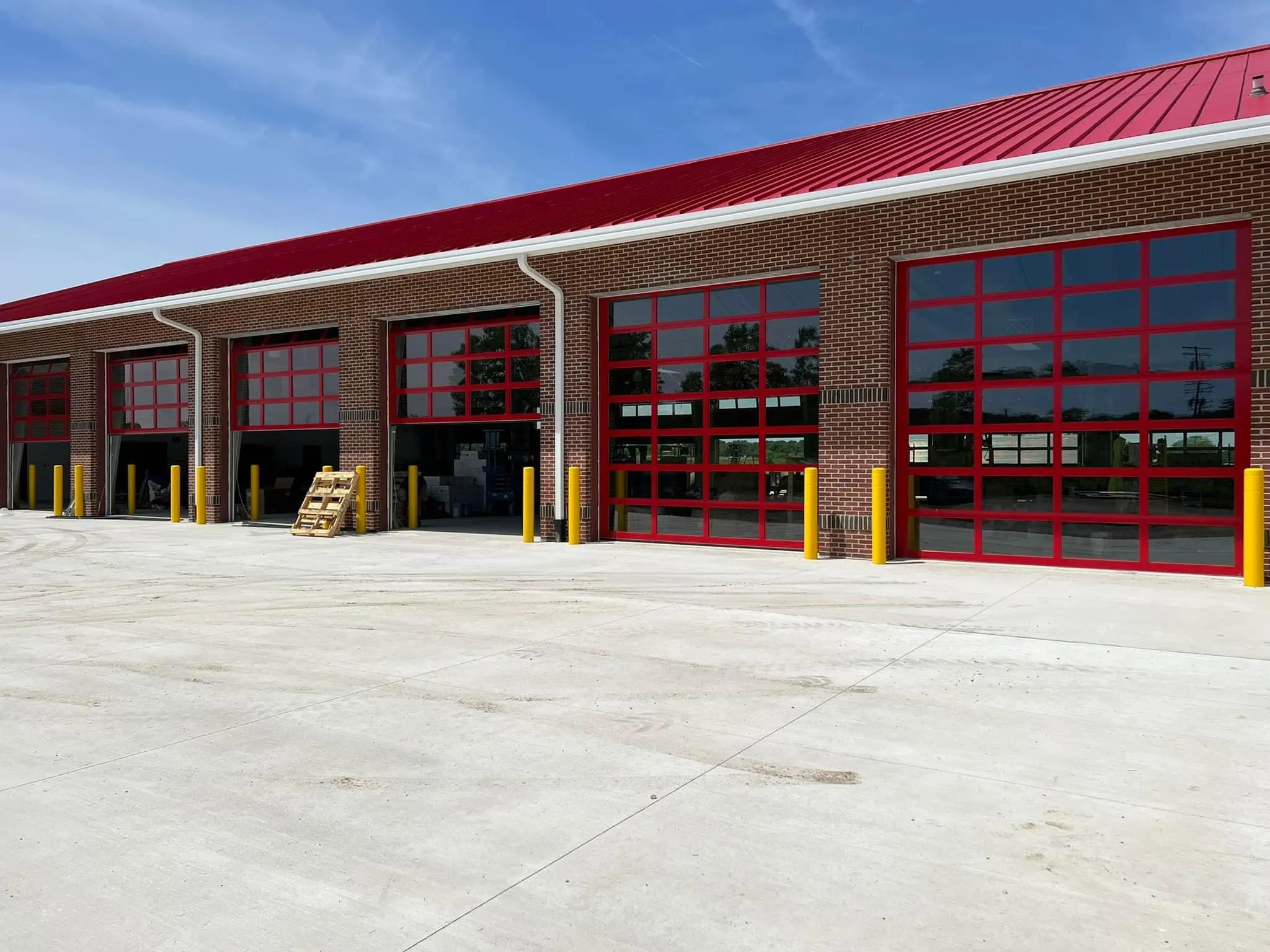 Long row of gray industrial overhead garage doors with small rectangular windows and yellow safety bollards on a wet concrete driveway.