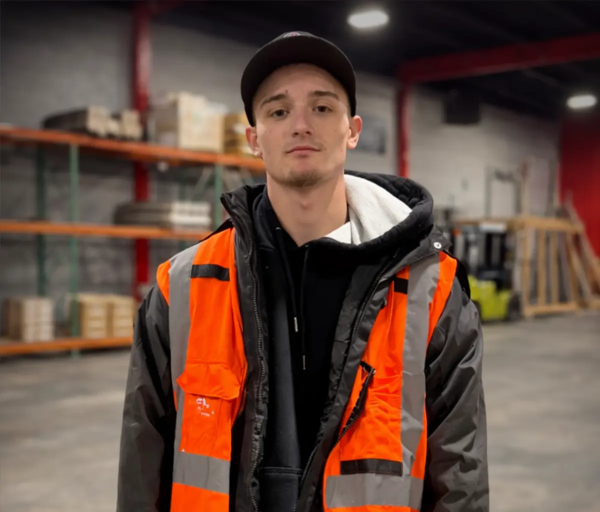 Young man wearing a black cap and orange safety vest over a black jacket in a warehouse setting.