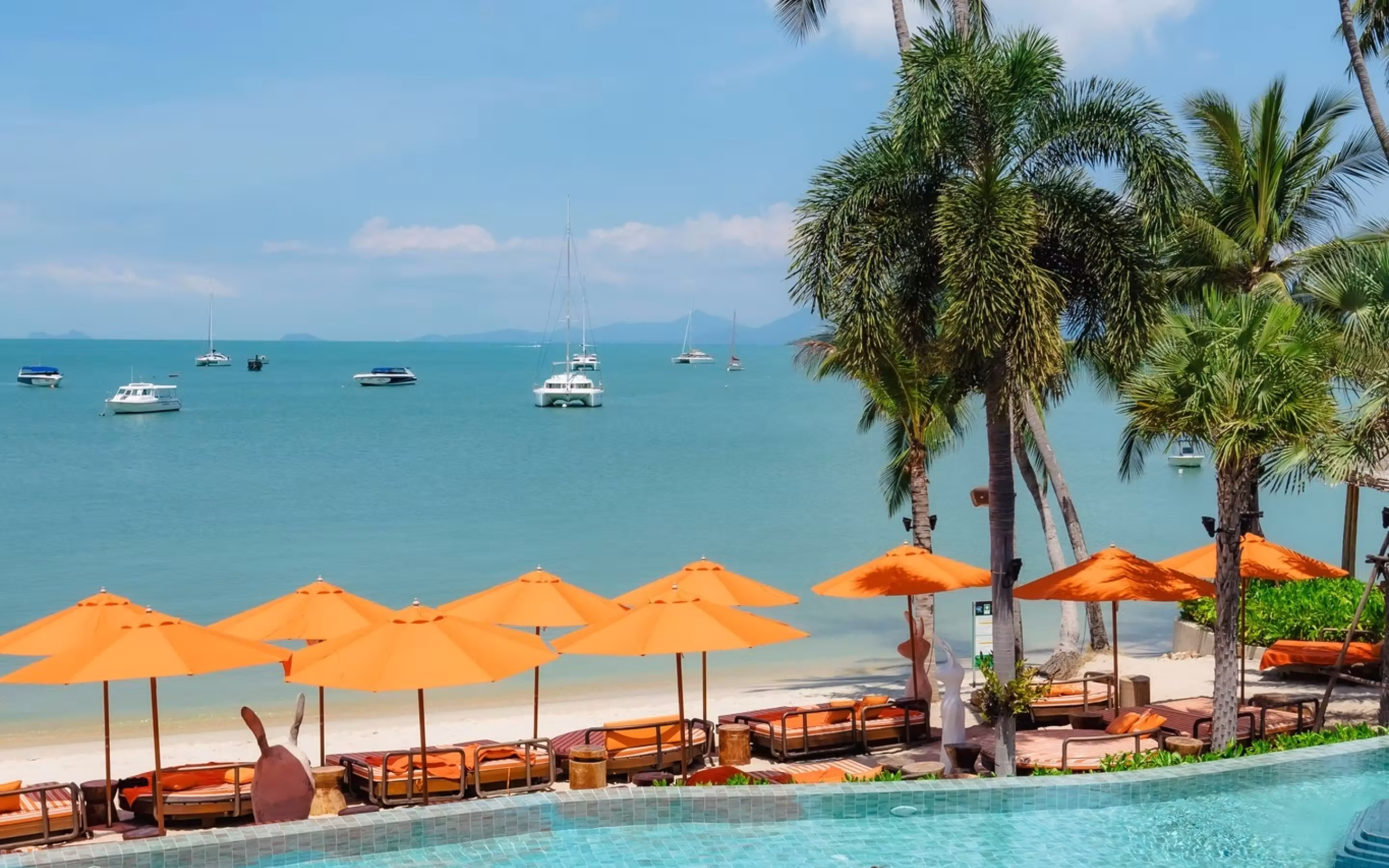 Beachfront scene with turquoise ocean, several anchored white boats, orange umbrellas, lounge chairs, and palm trees under a clear blue sky.