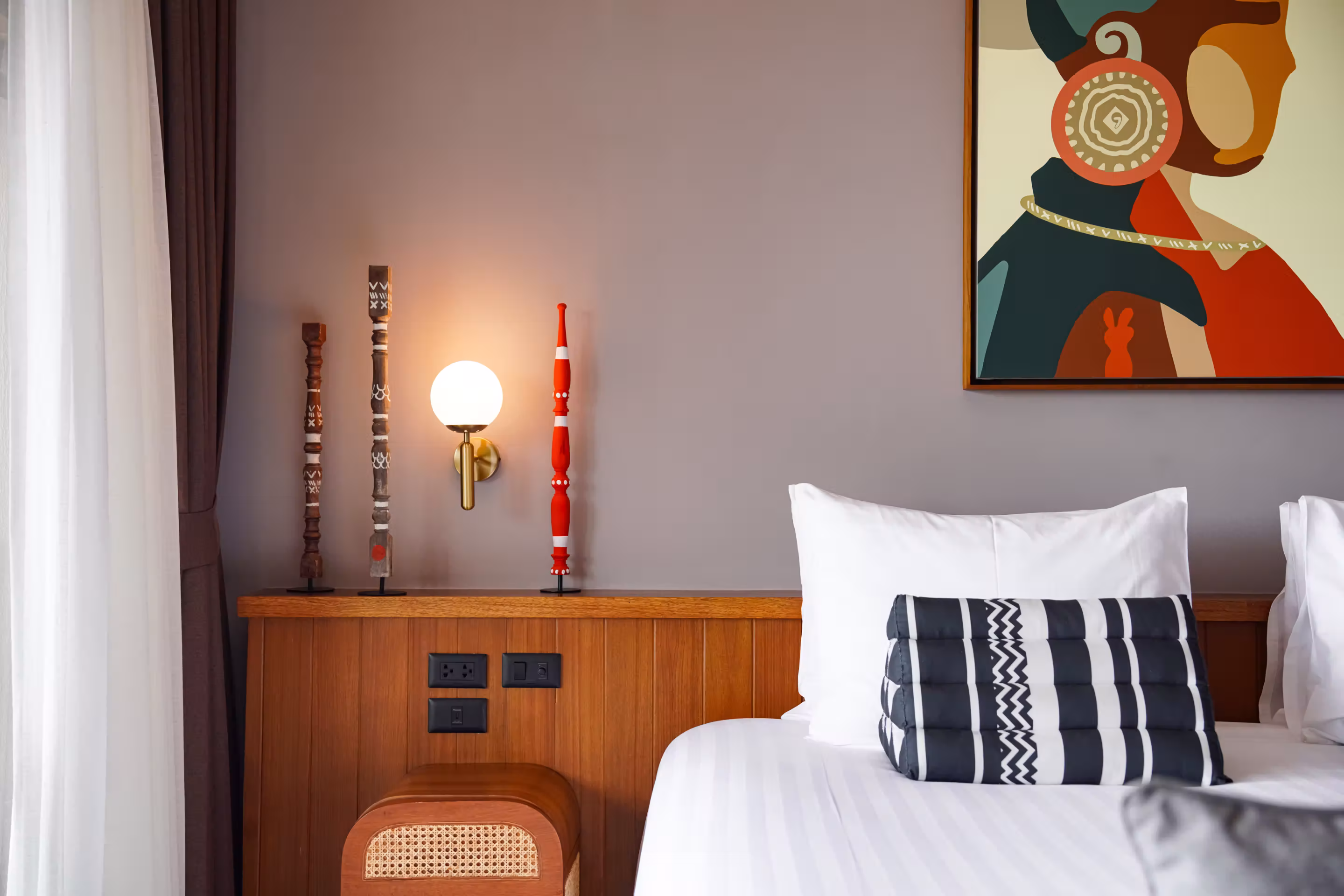 Bedroom with a bed featuring white pillows and black-and-white patterned triangular cushions, a wooden chair with an orange pillow, and abstract wall art above.