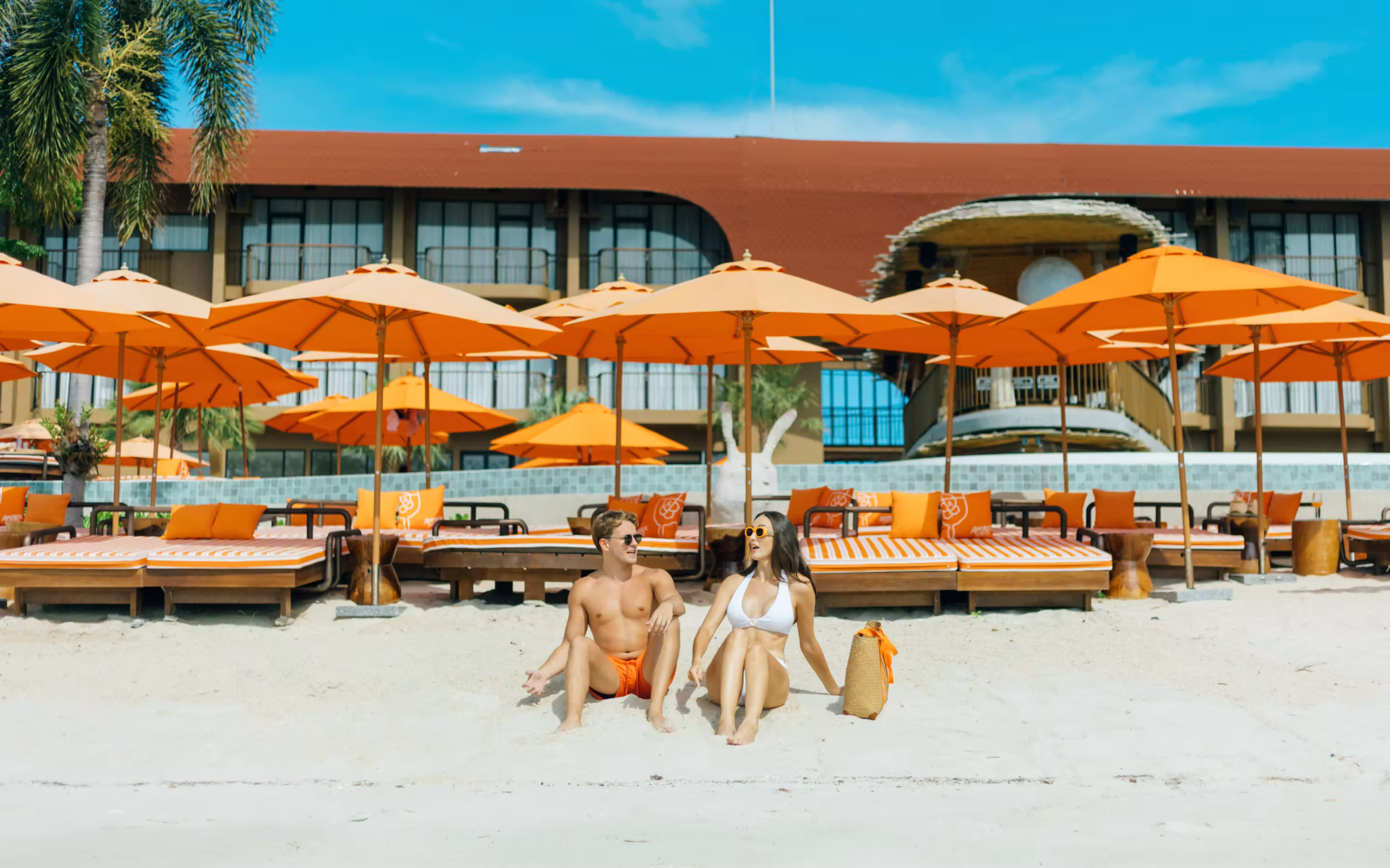 Tropical resort pool with orange umbrellas and lounge chairs, palm trees, and ocean view with boats.