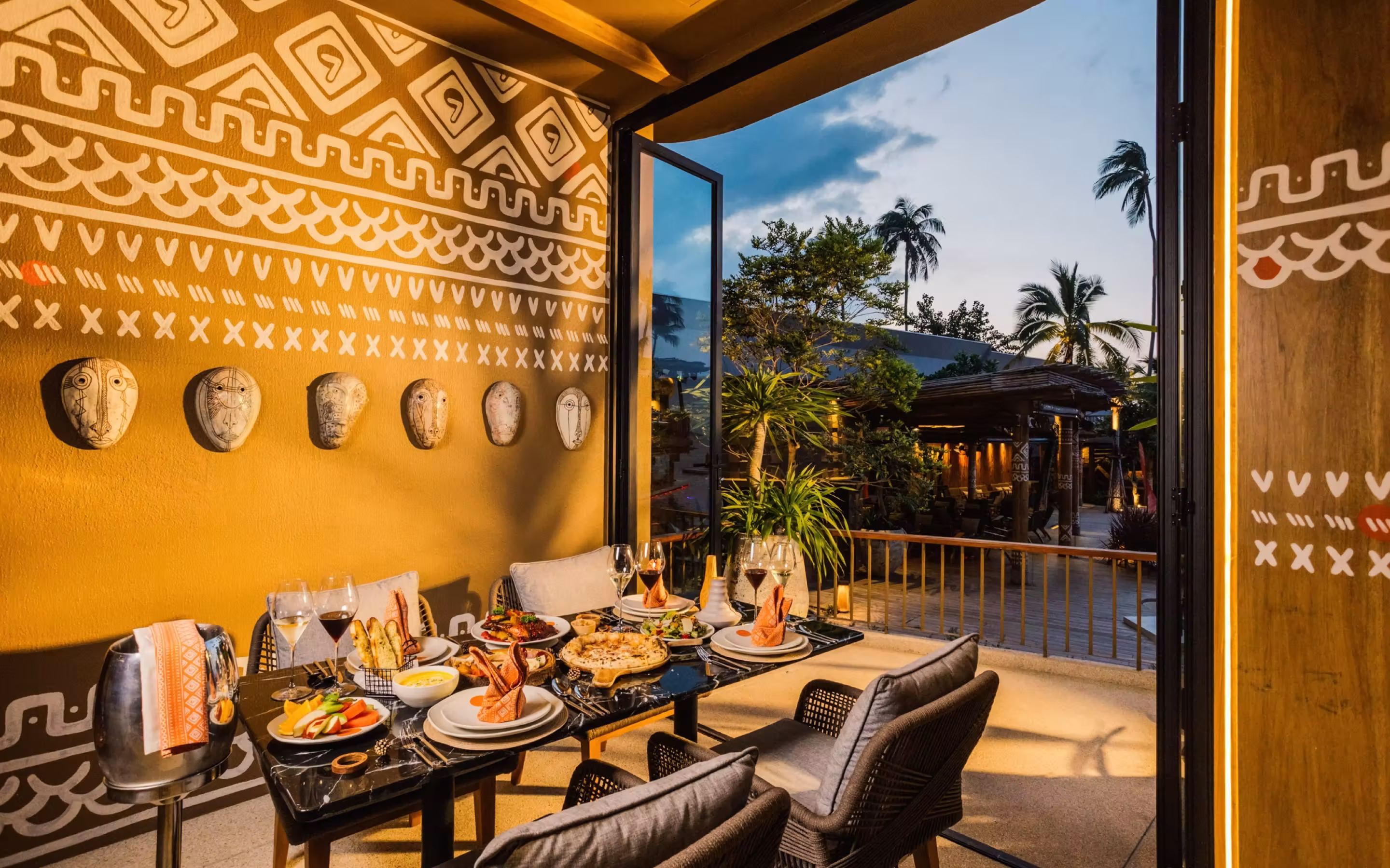 Outdoor dining area with a black marble table set for four, featuring plates with folded orange napkins, wine glasses, assorted dishes, and a backdrop of a yellow wall decorated with tribal masks and white geometric patterns, overlooking palm trees at dusk.