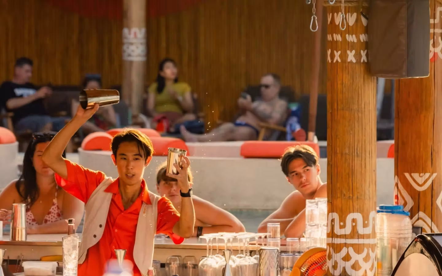 Bartender in an orange shirt shaking cocktail shakers behind a bar with patrons relaxing in the background at a tropical venue.