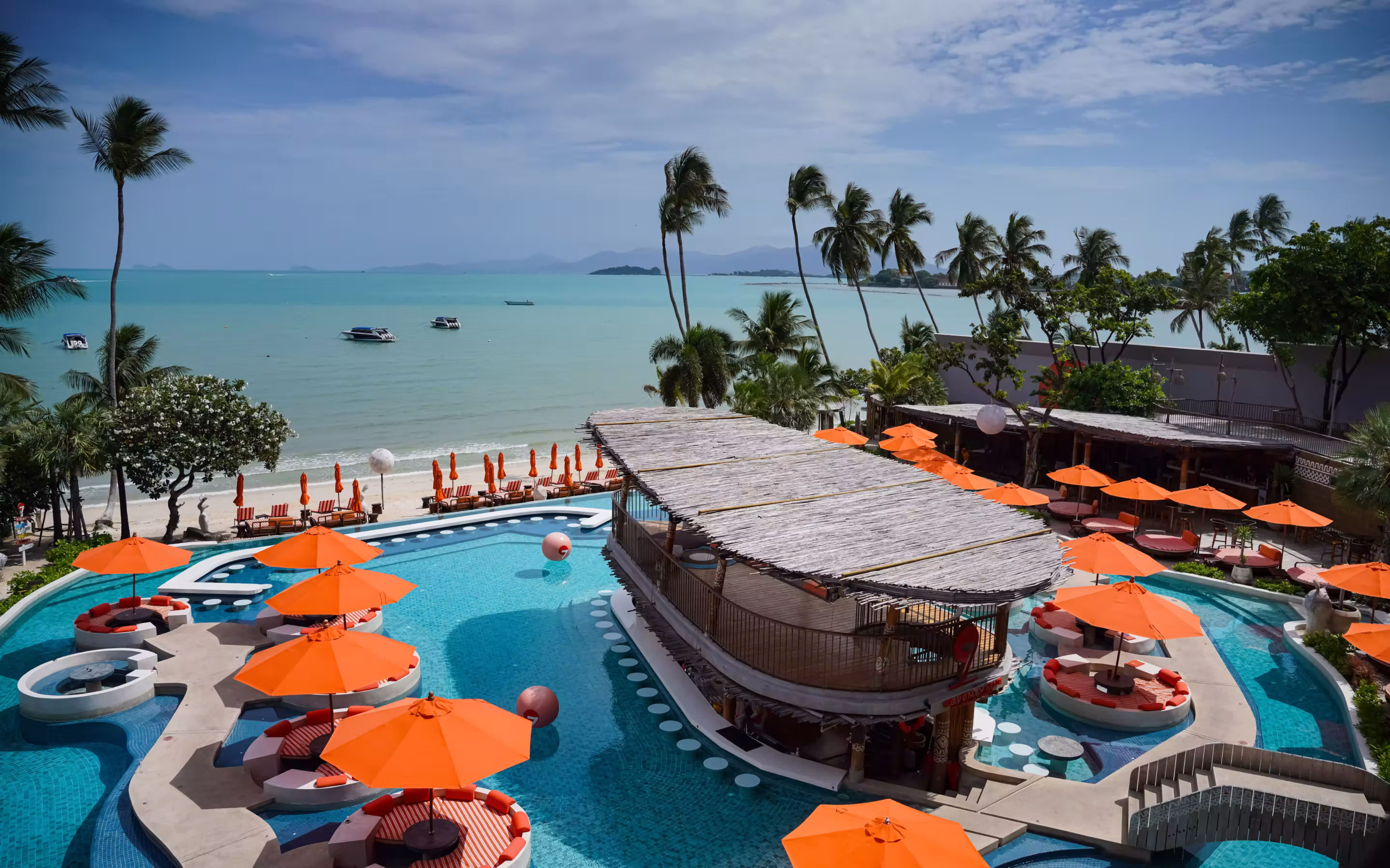 Resort pool area with orange-cushioned round loungers, large floating orange balls, a thatched roof bar, and a building with curved red roof under a clear blue sky.