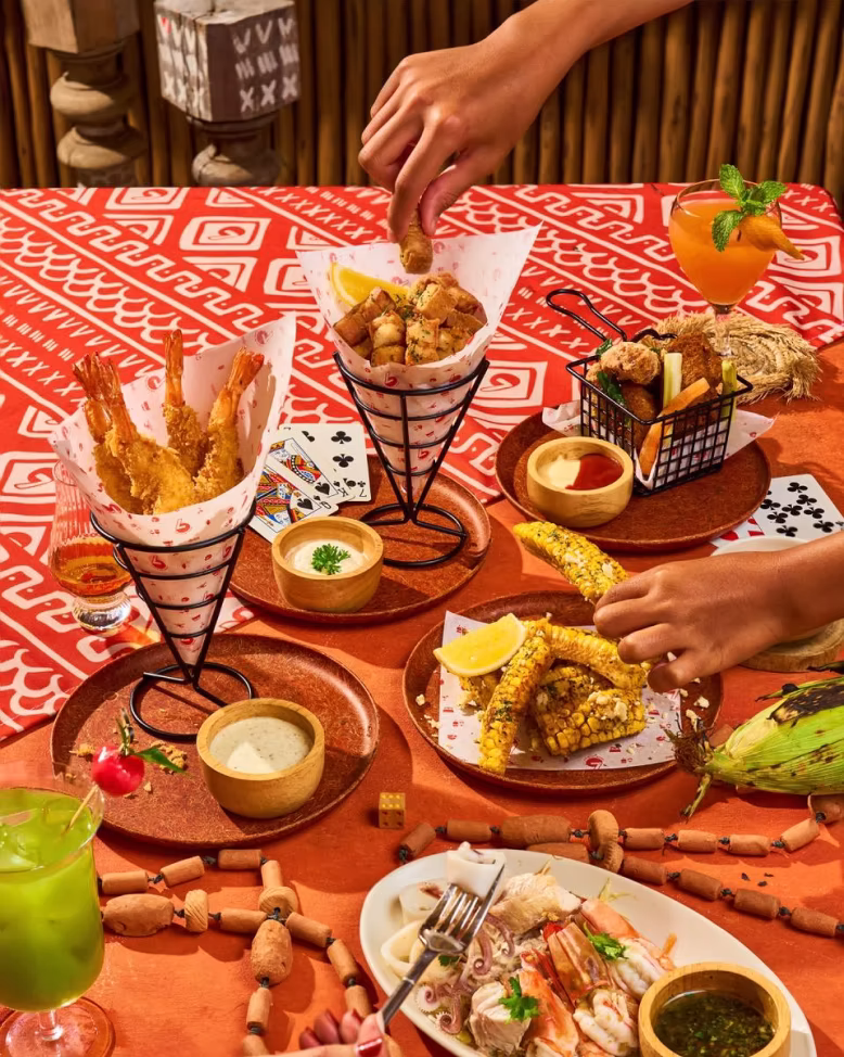 Table with assorted appetizers including fried shrimp, grilled corn, dipping sauces, seafood platter, and cocktails with hands reaching for food.