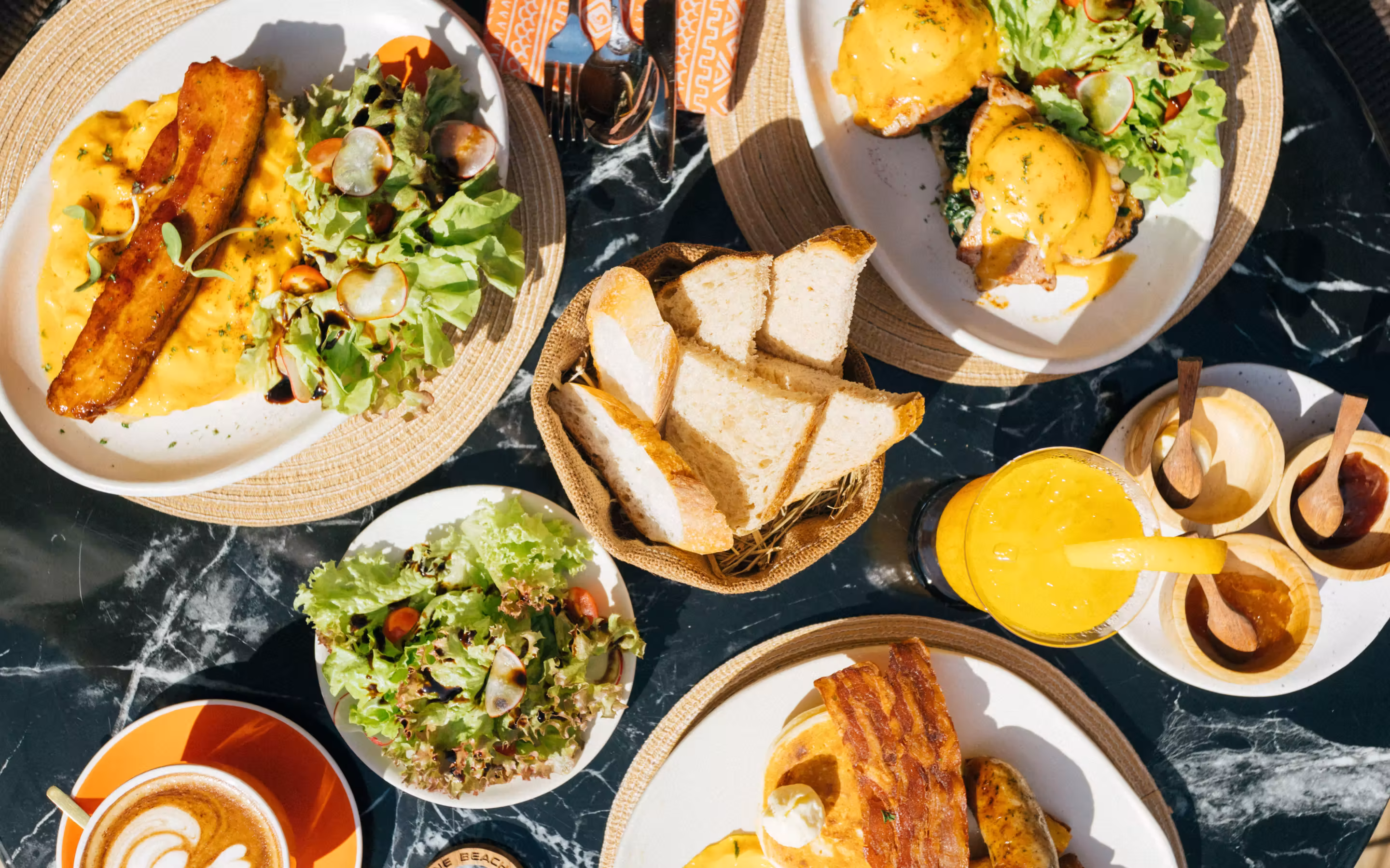 Table set with plates of breakfast including scrambled eggs with bacon, eggs Benedict with greens, fresh salad, sliced bread, orange juice, and a cup of coffee with latte art.