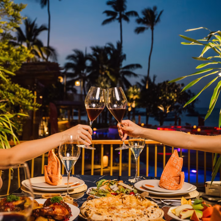 Two people clinking glasses of red wine over a table set with pizza and other dishes at an outdoor evening dining area with palm trees in the background.