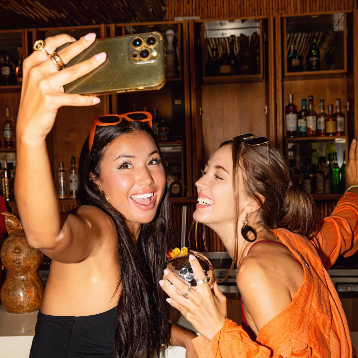 Two young women smiling and taking a selfie together in a bar, one holding a cocktail glass with garnish.