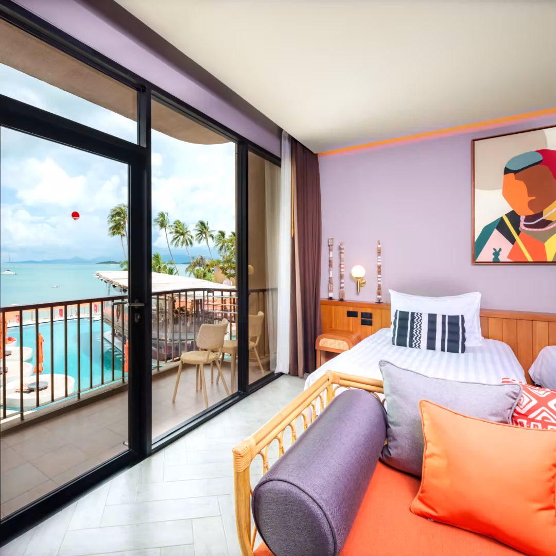 Modern hotel room with rattan sofa, wooden tables, desk, and balcony overlooking palm trees and ocean.