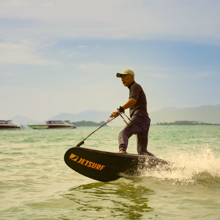 Man wearing a cap riding a JetSurf electric surfboard over water with boats and mountains in the background.