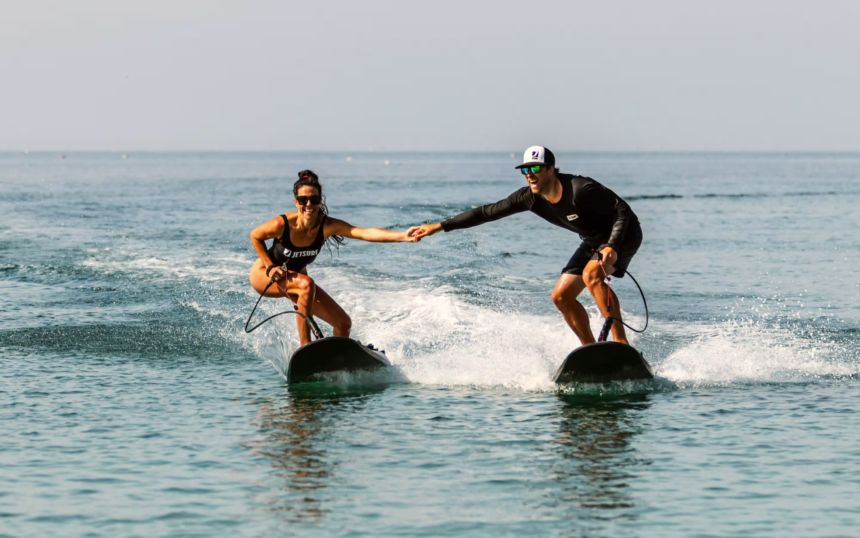 A man and a woman in sunglasses holding hands while riding electric surfboards on calm ocean water.