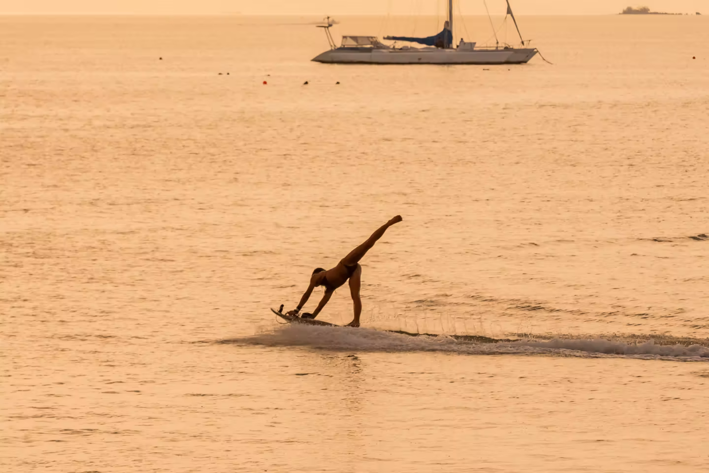 Person balancing on an electric surfboard on calm water during sunset with a sailboat in the background.