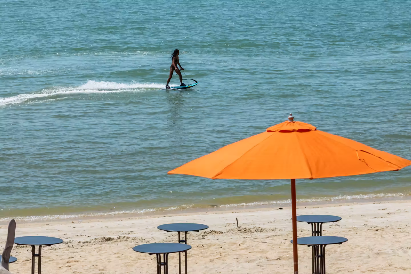 Person riding an electric surfboard on calm ocean water near a sandy beach with empty tables and an orange beach umbrella.