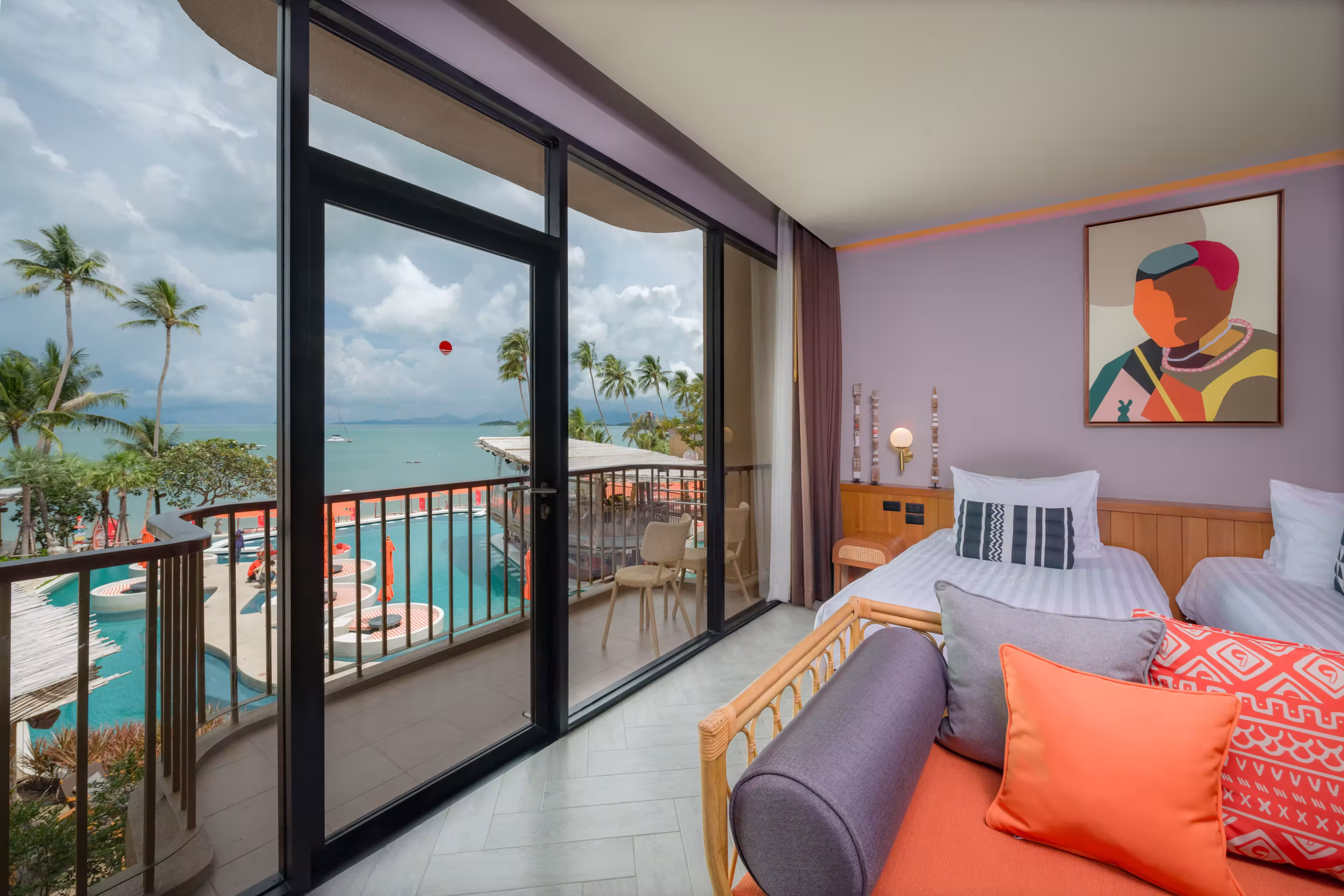 Hotel room with two beds, colorful pillows, abstract wall art, and a balcony overlooking a pool and ocean with palm trees.