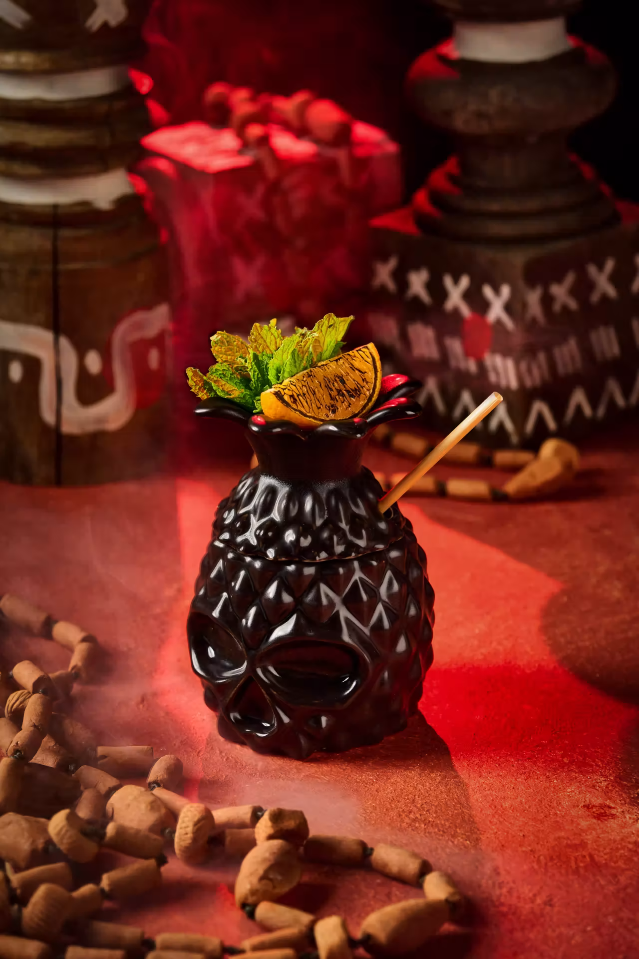 Black skull-shaped drink container with textured surface, garnished with mint and grilled orange slice, set on a red surface with decorative beads and wooden carvings in the background.