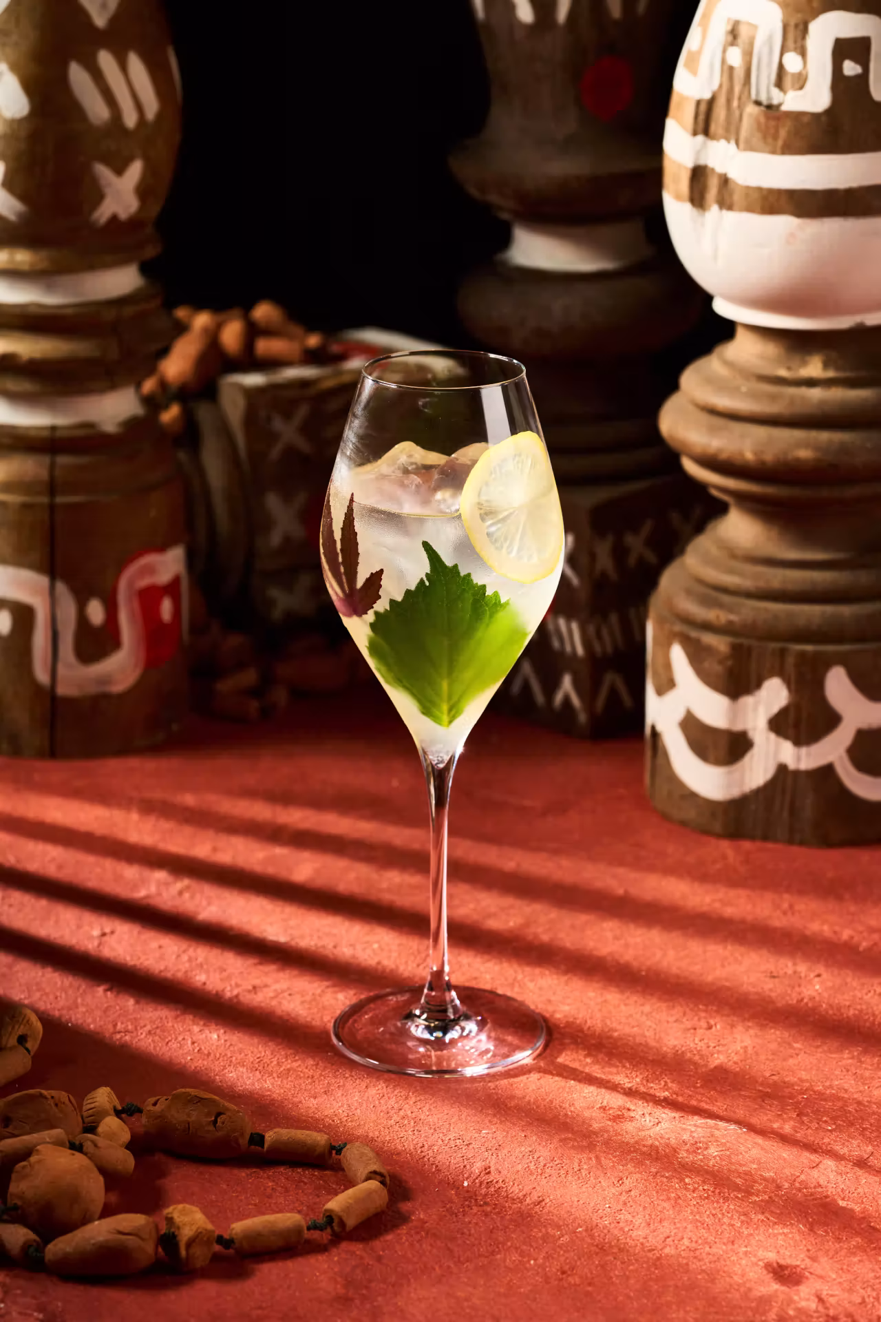 Wine glass with lemon slice, green and purple leaves, and ice cubes on a reddish surface with wooden ornamental objects in the background.