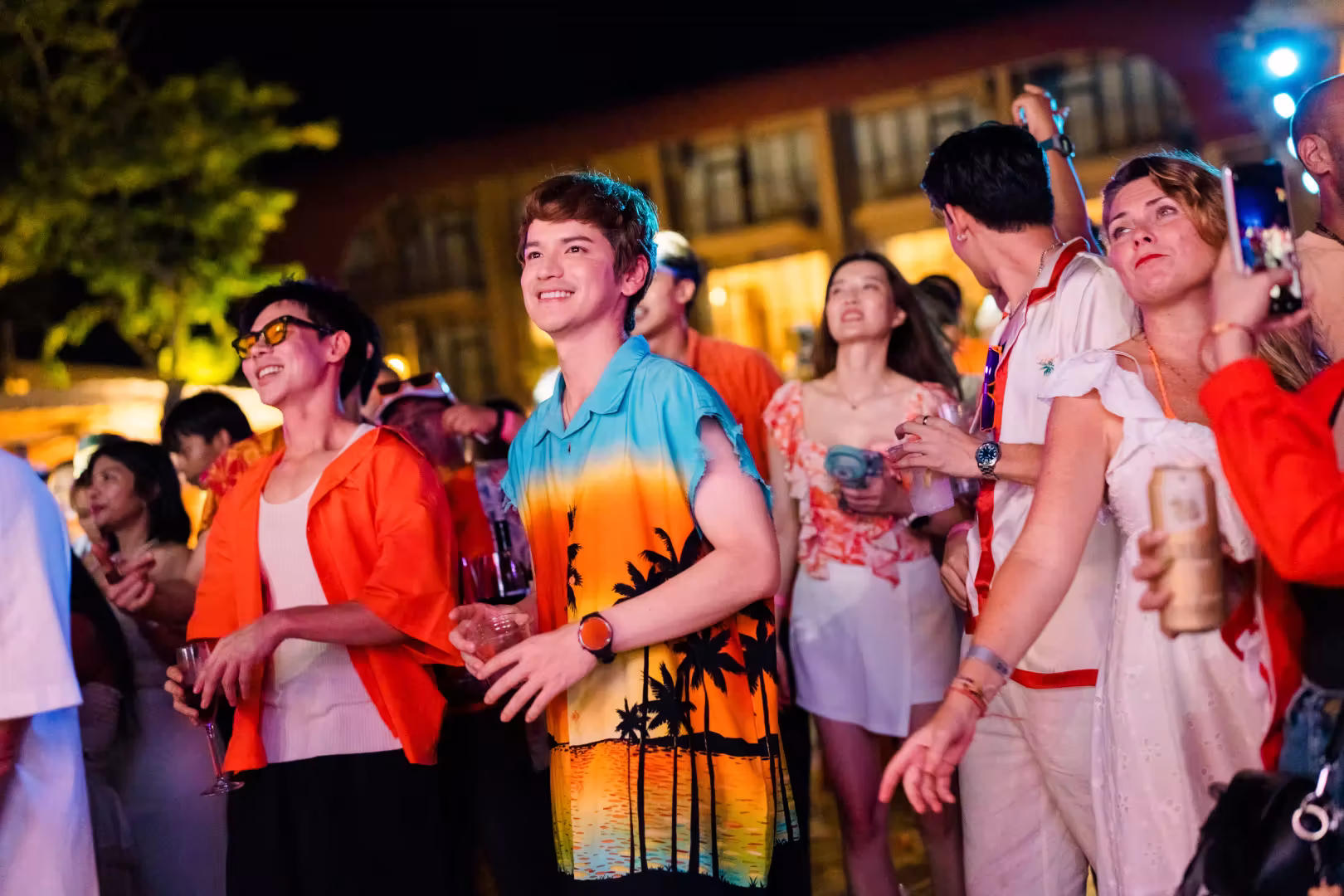 Group of young adults enjoying a lively outdoor night party, holding drinks and smiling.
