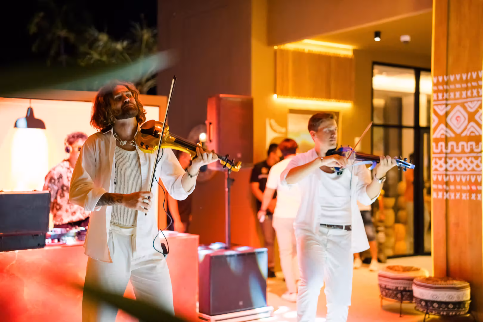 Two men playing violins at a nighttime indoor-outdoor event with warm lighting.